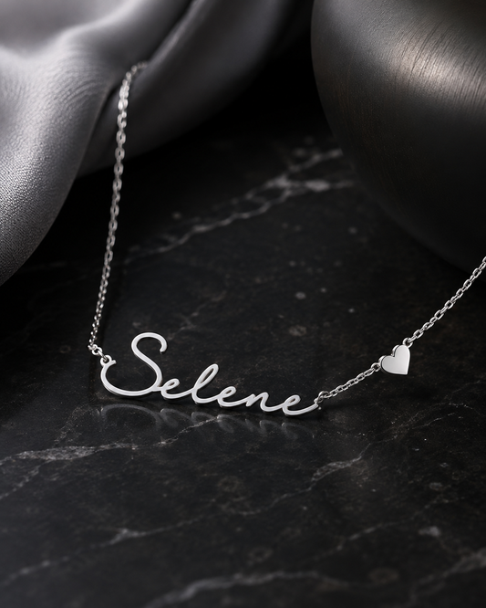 A silver name necklace spelling 'Selene' in cursive script, displayed on a dark marble surface with draped satin fabric and a dark metallic sphere in the background.