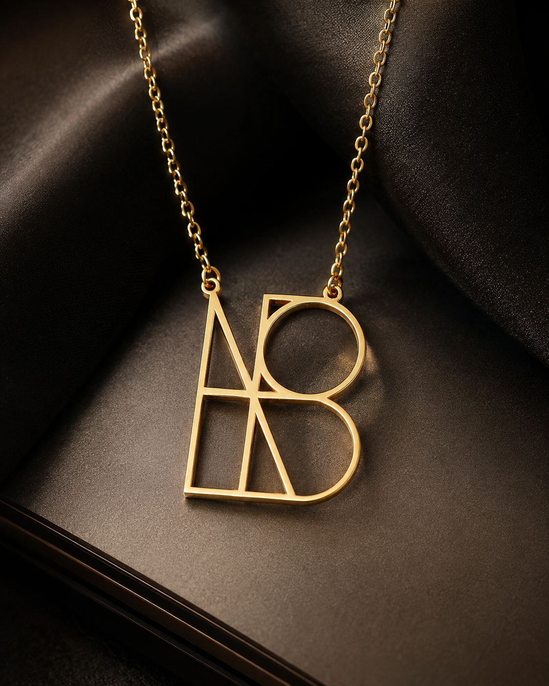 A gold initial necklace featuring interlocking stylized letters 'A' and 'B' on a dark satin fabric background.