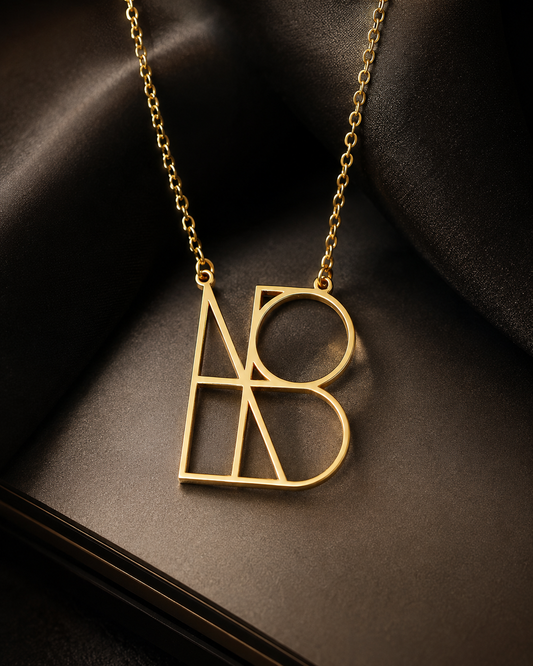 A gold initial necklace featuring interlocking stylized letters 'A' and 'B' on a dark satin fabric background.